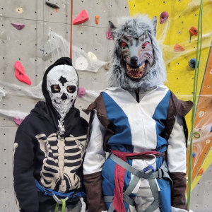 Deux personnes costumées devant un mur d'escalade - l'une portant un costume de squelette et l'autre un masque de loup avec des vêtements bleus et blancs. Les deux portent des baudriers d'escalade.